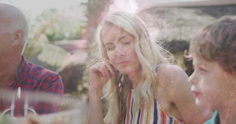 Contemplative woman sitting at sunlit garden table during family outdoor lunch gathering