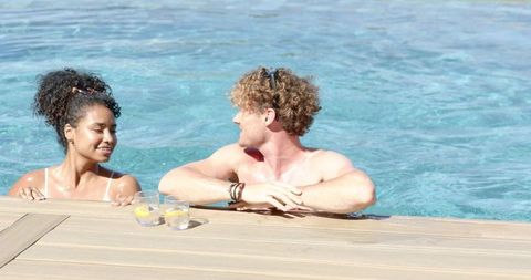 Interracial couple relaxing at resort pool edge with lemon drinks on wood deck, sunlit