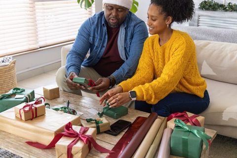 African american couple wrapping holiday gifts at home, tying ribbons, smiling in sunlit living room