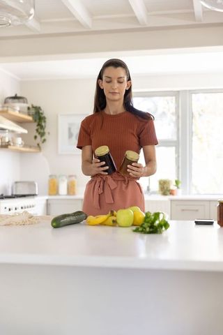 Mid-adult woman comparing jarred preserves while preparing fruit on sunlit kitchen island