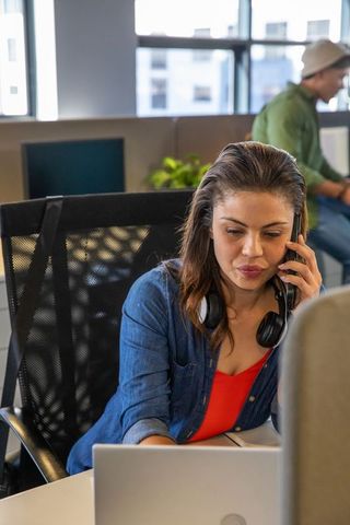 Young professional woman working on laptop while talking on smartphone in modern open-plan office