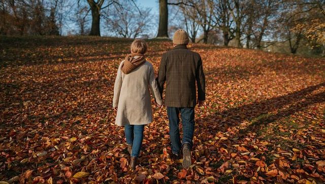 Senior couple holding hands walking through autumn park covered in fallen leaves at golden hour