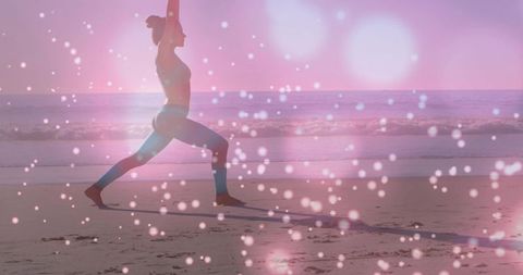 Woman practicing yoga lunge on beach, raising arms amid pink bokeh sunrise for wellness