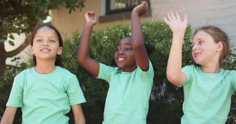 Cheering school-age girls raising arms in backyard wearing matching light green shirts