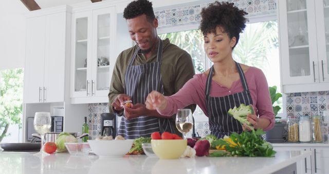 Happy African American couple enjoying cooking and drinking wine in a modern kitchen. This image might be used to promote the idea of quality time spent at home, kitchen-related products, relationship and family-oriented content, lifestyle blogs, romantic getaways, or advertisements focusing on modern living and love.