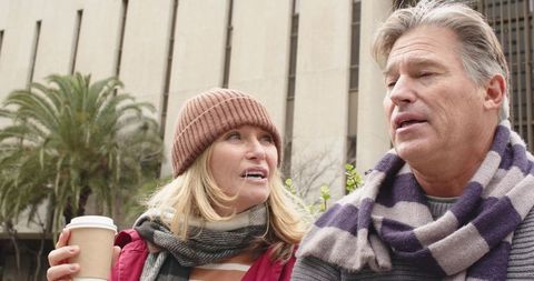 Mature couple chatting on urban bench holding takeaway coffee wearing scarves and beanie