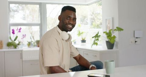 Smiling man tapping smartphone at kitchen counter wearing headphones minimalist sunlit