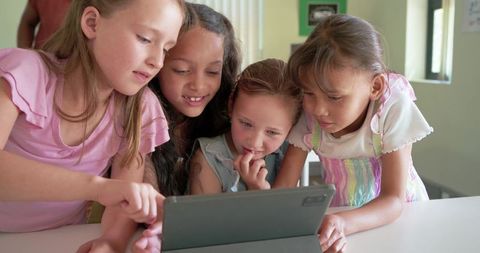 Diverse schoolgirls leaning over tablet, collaborating and learning together in classroom