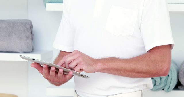 Person holding digital tablet in a minimalist and clean home interior, depicted wearing a white shirt, emphasizing a modern and tech-savvy lifestyle. This image is ideal for advertisements and articles related to contemporary living, digital habits, and technological devices. It can also serve well in subjects focusing on home organization, interior design, or personal workspace setups.