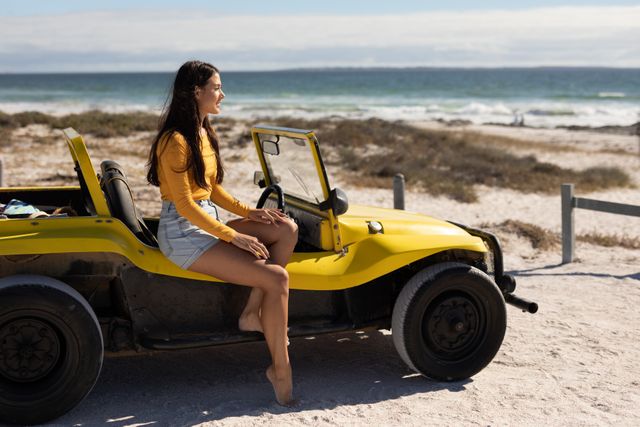 Woman sitting on a yellow beach buggy, smiling and admiring the sea view on a sunny day. Ideal for use in travel blogs, vacation advertisements, and lifestyle magazines promoting summer adventures and beach holidays.