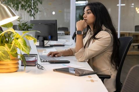 Indian woman working at modern open-plan office desk with laptop, plant and smartwatch