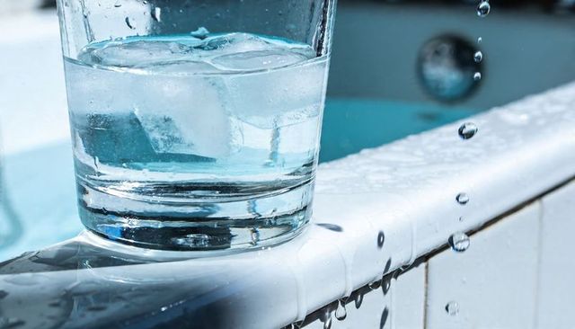 Clear glass tumbler with ice water on pool edge, dripping droplets and sunlit reflections