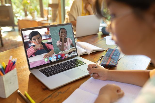 Asian teenage girl writing notes while studying online through video call on laptop at home. Unaltered, screen, internet, wireless technology, education, student and e-learning concept.