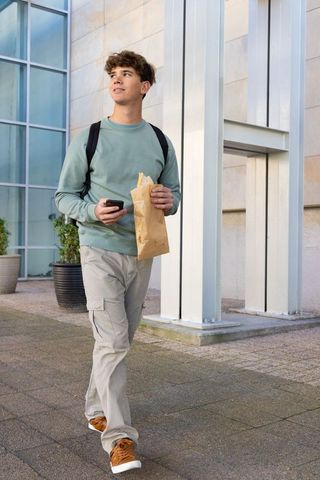 Young man walking with backpack and paper lunch bag checking smartphone near glass facade