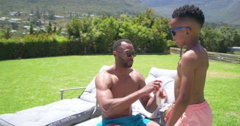 African american father applying sunscreen to son on outdoor lounger sunny backyard day