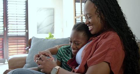 African american mother and daughter laughing sharing smartphone on sunlit sofa