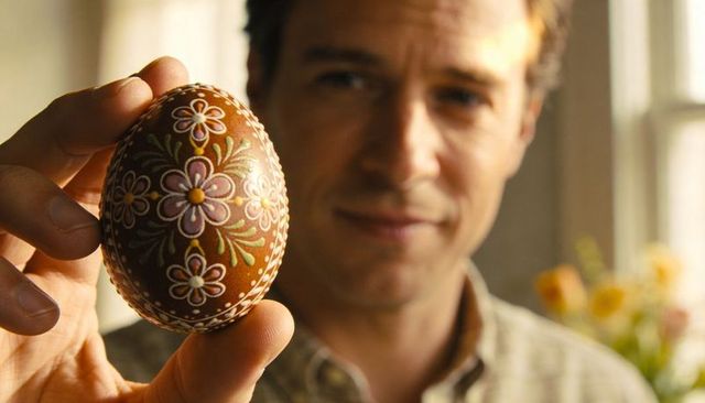 Man holding ornate brown pysanka egg with hand-painted floral motif by sunlit window