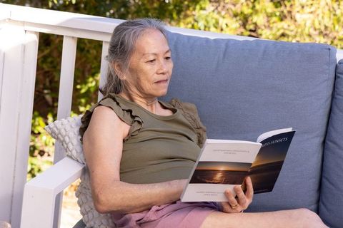 Senior asian woman relaxing on sunlit porch sofa reading paperback, outdoor leisure moment