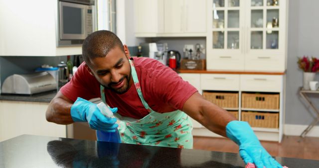 Man wearing apron and gloves, engaged in cleaning kitchen counter with spray bottle, conveying positive attitude towards household chores. Suitable for articles on cleaning tips, household chores, domestic life, or product advertisements for cleaning supplies.
