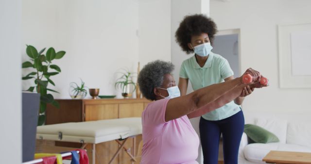 Senior woman lifting weights with assistance from physical therapist in a home environment. Ideal for content about senior wellness, at-home physical therapy, elderly fitness routines, and healthcare guidance. Great for promoting personal training services, rehabilitation programs, and elderly care information.