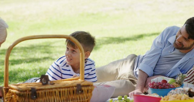 A young Caucasian boy enjoys a sunny day at a picnic with a middle-aged man, his father, with copy space. They are relaxing on the grass, surrounded by a basket and fresh fruits, sharing a moment of leisure and bonding.