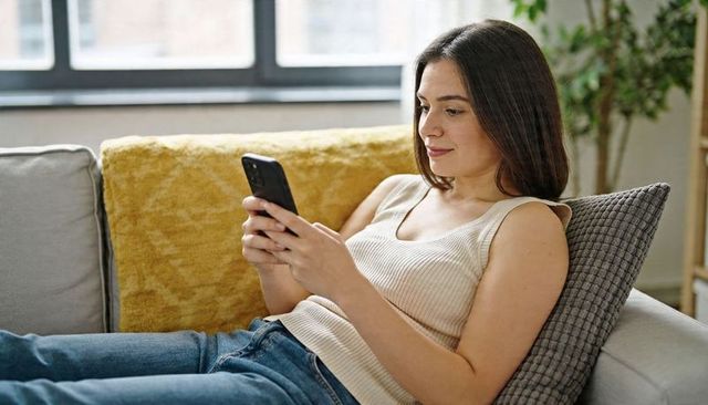 Young woman relaxing on sofa scrolling smartphone in bright minimalist living room
