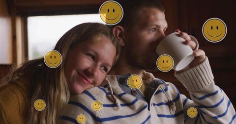 Couple cuddling in knit sweaters sipping coffee with smiley stickers in cozy cabin lounge