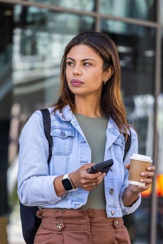 African american commuter holding smartphone and coffee by modern glass building