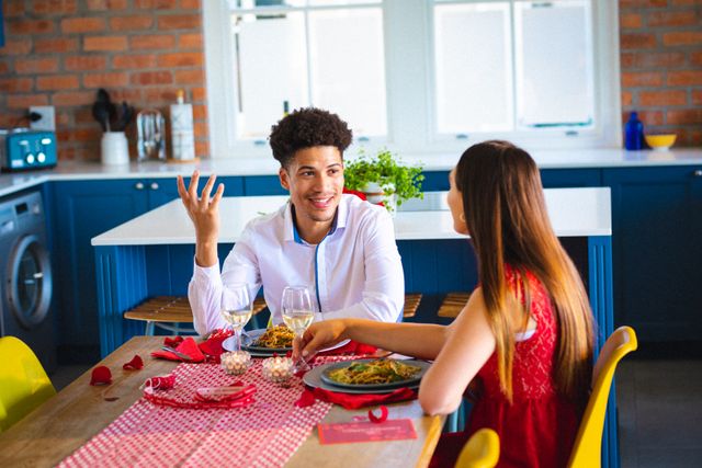 Young couple enjoying a romantic lunch date in a modern kitchen. The man is talking and smiling while the woman listens attentively. The table is set with food, wine, and candles, creating a warm and intimate atmosphere. This image is perfect for use in lifestyle blogs, relationship advice articles, or advertisements promoting home dining experiences.