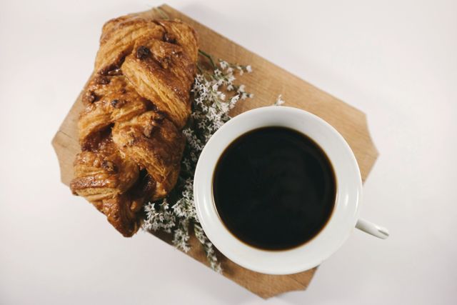 Cup of black coffee placed next to sweet Danish pastry on wooden board. Floral decoration beside coffee adding touch of elegance. Ideal for use in breakfast promotions, bakery adverts, and coffee shop menus.
