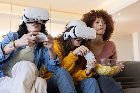 Multicultural Friends Playing VR on Couch, Sharing Snacks, Wearing White Headsets