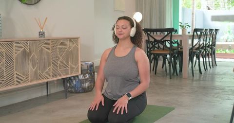 African american woman meditating on yoga mat at home wearing headphones and smartwatch
