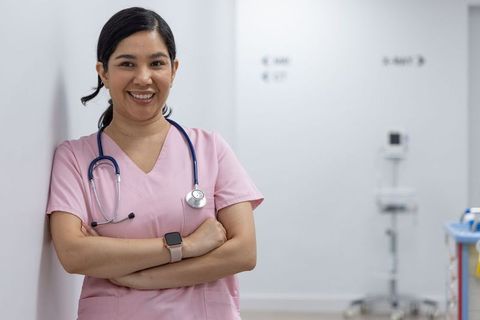 Smiling Nurse Wearing Pink Scrubs and Stethoscope Leaning in Clinic Corridor with Smartwatch