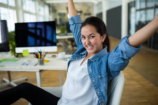 Young female graphic designer celebrating success at her desk in a modern office. Ideal for use in articles about workplace happiness, creative professions, business success, and office culture. Can also be used in promotional materials for design agencies or career development programs.