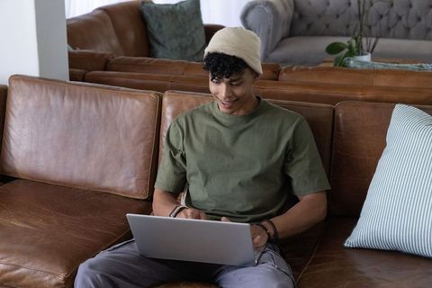 African man working on laptop on leather sofa wearing beanie and bracelets, relaxed