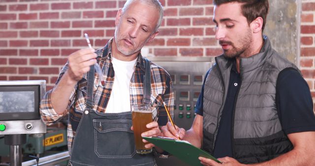Brewing expert and assistant closely examining a glass of beer and taking notes on clipboard. Ideal for use in articles and advertisements related to craft beer production, quality control in breweries, teamwork in manufacturing processes, and the craft beverage industry.