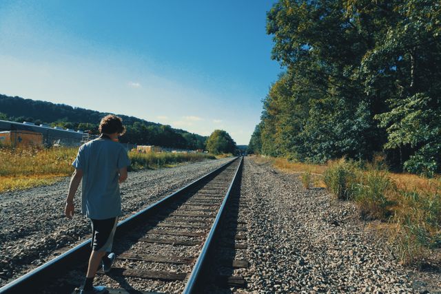 Teenage boy casually walking along railroad tracks surrounded by lush greenery under clear blue sky in countryside. Ideal for themes of adventure, exploration, serenity, and rural life. Perfect for travel blogs, adventure inspiration, or lifestyle publications highlighting youthful freedom and connection with nature.