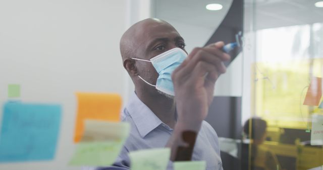 Businessman wearing mask and writing on glass wall surrounded by sticky notes. Perfect for illustrating modern office environments, team collaboration, project planning, and adapting to new workplace health protocols.
