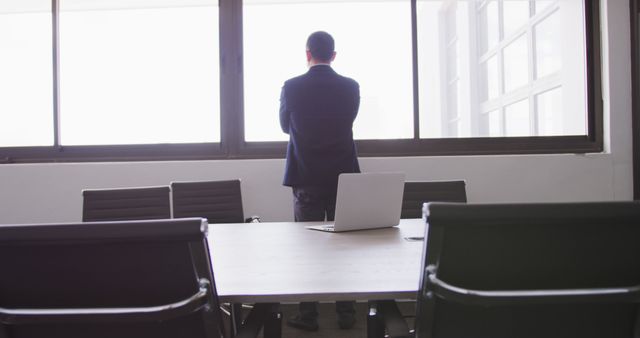 Businessman standing alone in conference room looking out window, creating a thoughtful and reflective atmosphere. Useful for concepts related to business, solitude, decision making, leadership, and office environments. Ideal for illustrating business articles, leadership training materials, and corporate presentations.