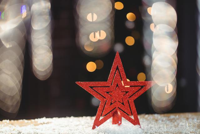 Red star ornament resting on artificial snow with blurred bokeh lights in the background. Ideal for holiday greeting cards, festive advertisements, Christmas-themed social media posts, and seasonal decorations.