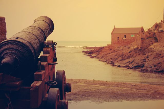 Historic iron cannon on wooden platform pointing towards calm sea with rugged shoreline. Stone building in background adds vintage appeal. Perfect for illustrating historical scenes, coastal defenses, or tranquil seaside locales.