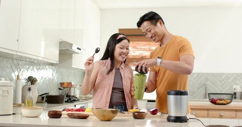 Asian couple preparing green smoothie in bright modern kitchen, smiling, healthy lifestyle