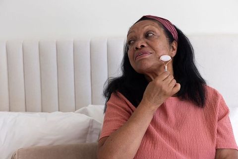 Senior african-american woman using rose quartz facial roller on jaw while sitting on bed