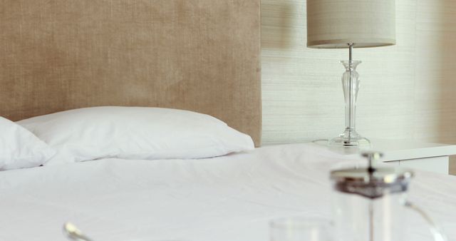 White linens on a bed with beige headboard and table lamp on nightstand suggesting a cozy and comfortable environment. Minimalist design evokes a peaceful atmosphere. Ideal for home decor blogs, lifestyle content, and hospitality marketing.