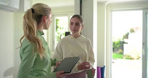 Female friends talking in bright home workout space holding tablet and water bottle