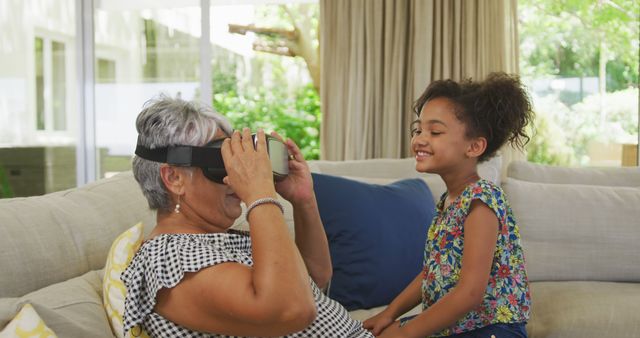 Grandmother enjoying virtual reality experience with her granddaughter watching with excitement. Perfect for use in advertisements about family bonding, modern technology, multigenerational activities, and tech-savvy seniors.