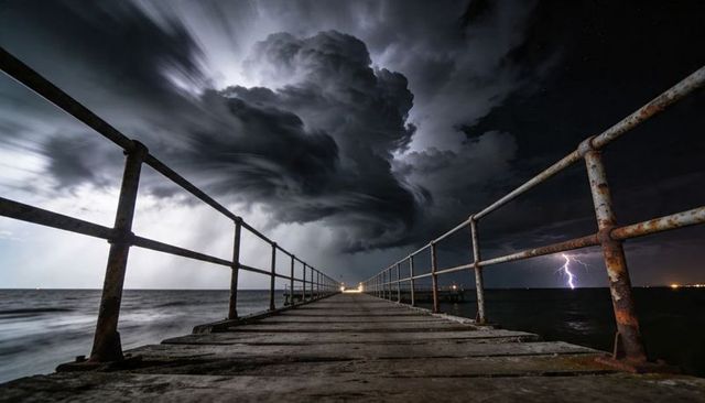 Stormy night pier leading to lightning and swirling thunderclouds over ocean horizon