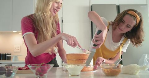Women decorating three-layer sponge cake on home kitchen island, smoothing frosting together