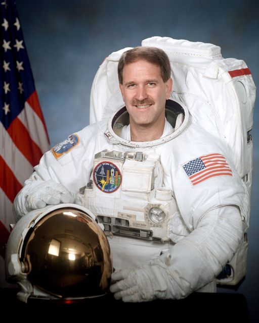 An astronaut in a space suit with the American flag in the background, holding a golden helmet and smiling at the camera. Ideal for use in educational materials, space exploration promotions, science exhibitions, and inspirational posters.