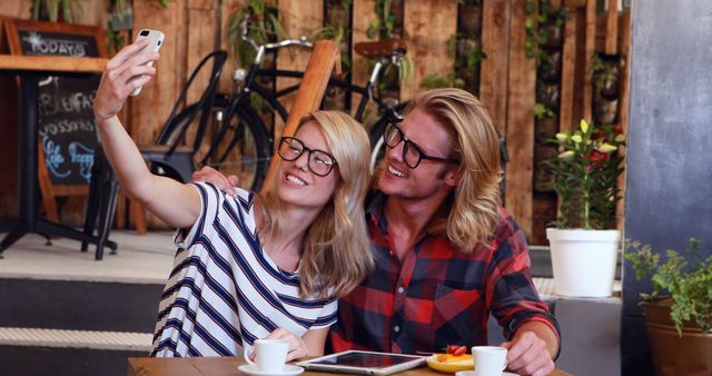 Young couple wearing glasses taking a selfie at a stylish cafe with a relaxed and cozy atmosphere. Ideal for themes about modern lifestyle, friendship, social media sharing, and young adult activities.